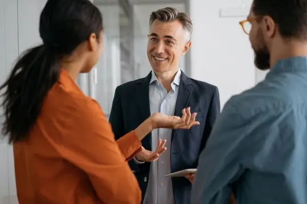 A group of three people in a business setting engage in a conversation. The career coach in the center holds a tablet and smiles, while the other two, a woman in a red shirt and a man in glasses, listen attentively.