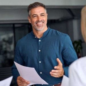 A smiling man in a blue shirt holds papers, engaged in conversation as a career coach. He stands in a modern office setting, interacting with colleagues, offering resume writing and career coaching services to guide their professional growth.