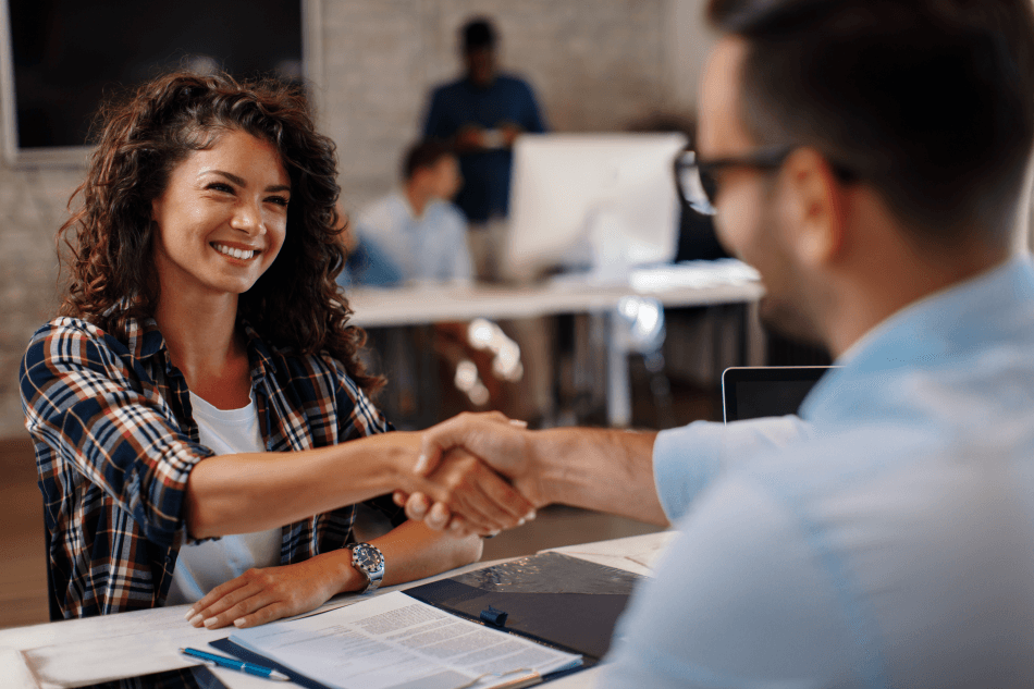 A smiling woman and a man shake hands across a desk in an office, with documents, a clipboard, and a Professional Resume in front of them. Other people work in the blurred background.